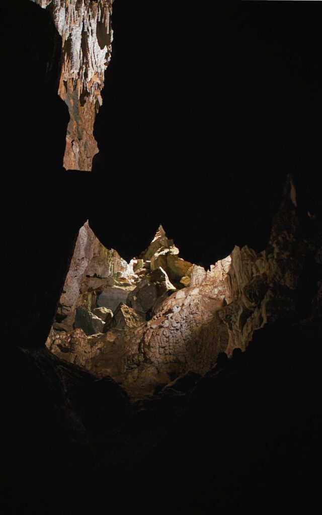 A fotografia captura o interior de uma caverna, onde formações rochosas são realçadas por iluminação. A luz enfatiza uma parte específica das rochas, projetando uma sombra que se assemelha a uma sala com lareira. As rochas circundantes apresentam formas irregulares e texturas diversas, incluindo espeleotemas. O escuro ao redor faz um contraste marcante com a área iluminada, destacando a silhueta projetada.