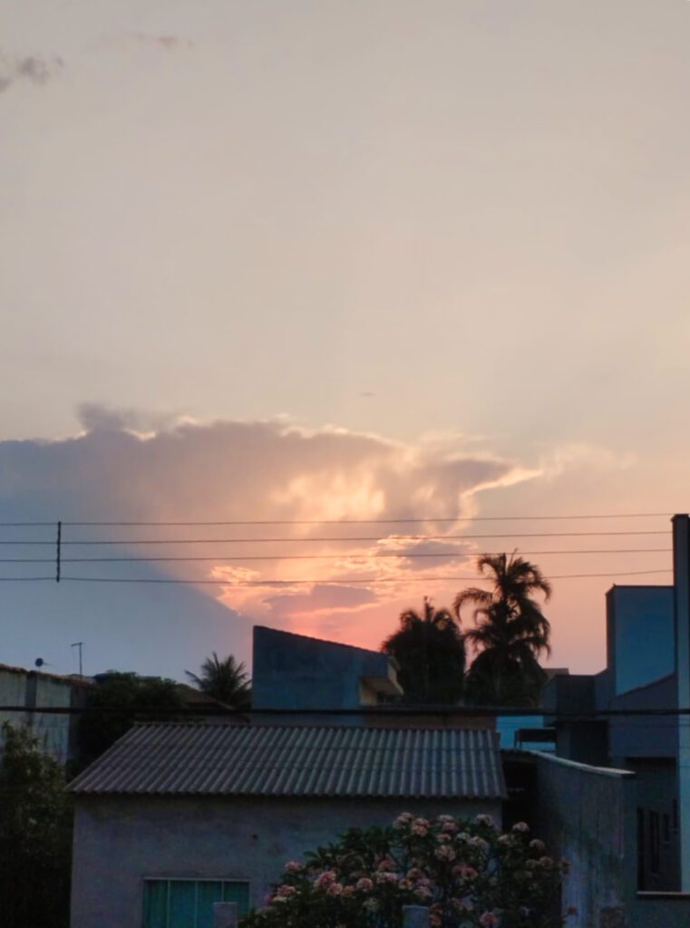 O céu se apresenta em um espetáculo de cores, com nuvens tingidas de laranja e rosa que se destacam contra o pano de fundo azul e cinza. A silhueta de palmeiras e estruturas urbanas compõe uma cena que harmoniza elementos naturais e construções humanas.